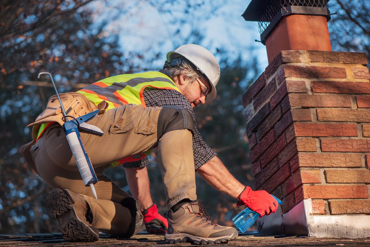 Roofer sprays water on brick chimney. Worker prepares cement application wearing white hard hat and safety gear